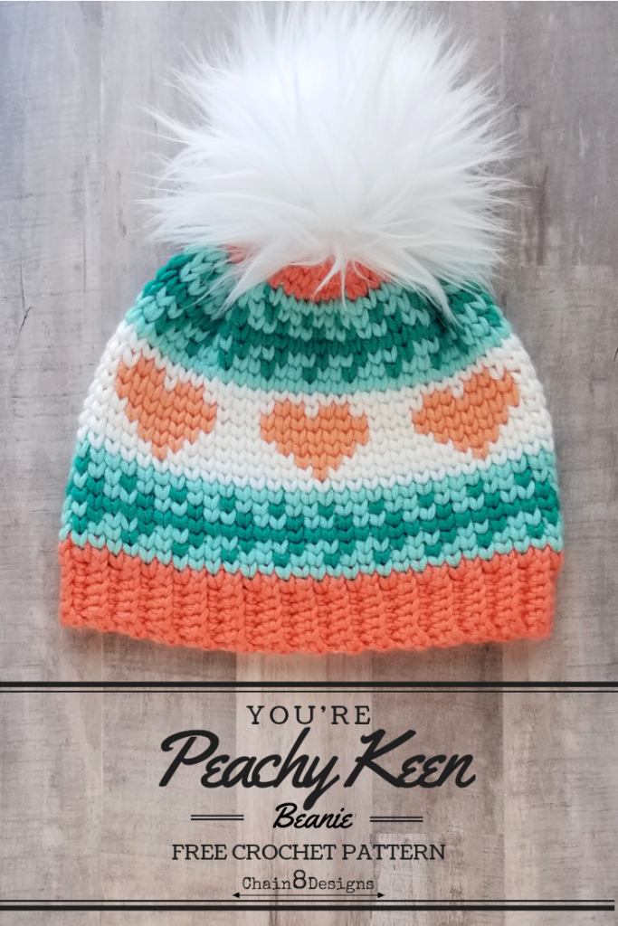 A crocheted beanie with peach hearts and a white fur pompom